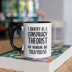 May include: White coffee mug with a black handle and black rim. The mug has the text "I identify as a conspiracy theorist. My pronouns are told/you/so" printed on it.