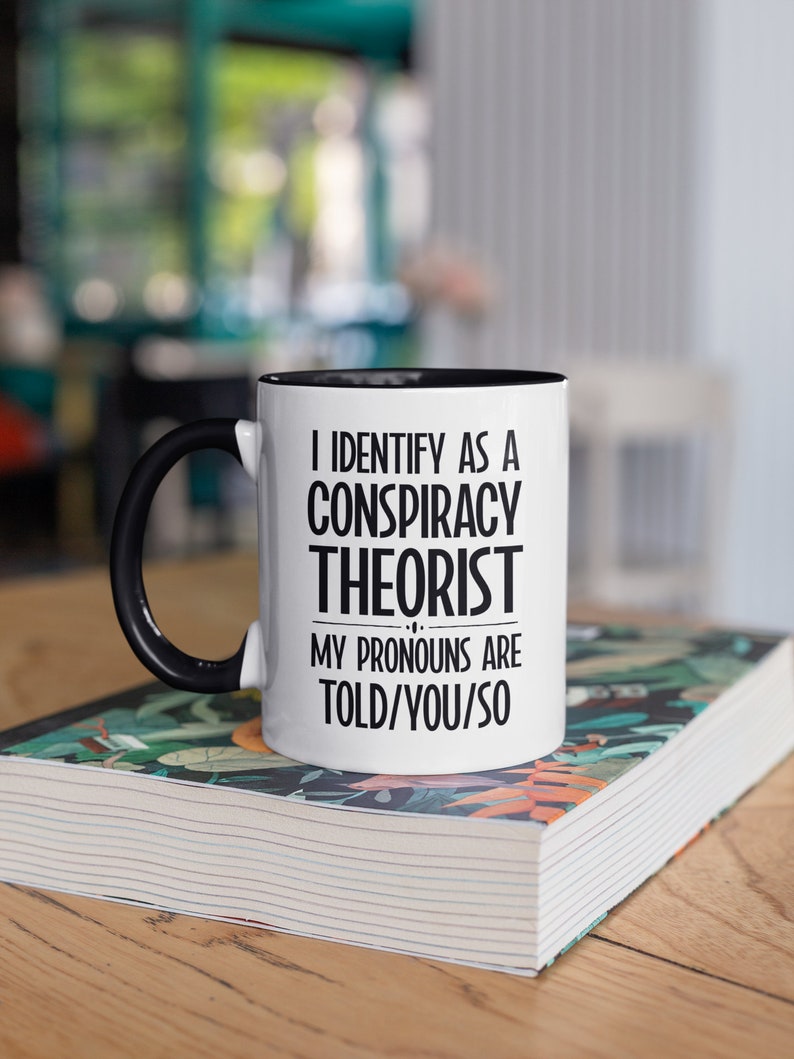 May include: White coffee mug with a black handle and black rim. The mug has the text "I identify as a conspiracy theorist. My pronouns are told/you/so" printed on it.