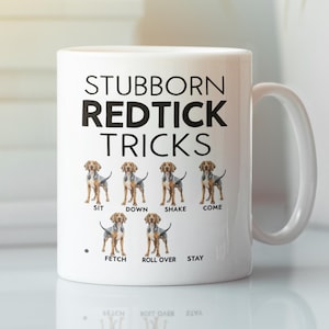 Pode incluir: Caneca de café de cerâmica branca com o texto "Stubborn Redtick Tricks" e ilustrações de Redtick Coonhounds realizando vários truques de cachorro, incluindo sentar, deitar, sacudir, vir, buscar, rolar e ficar.