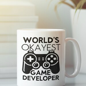 May include: White ceramic mug with a black design that says "World's Okayest Game Developer" with a black video game controller graphic.