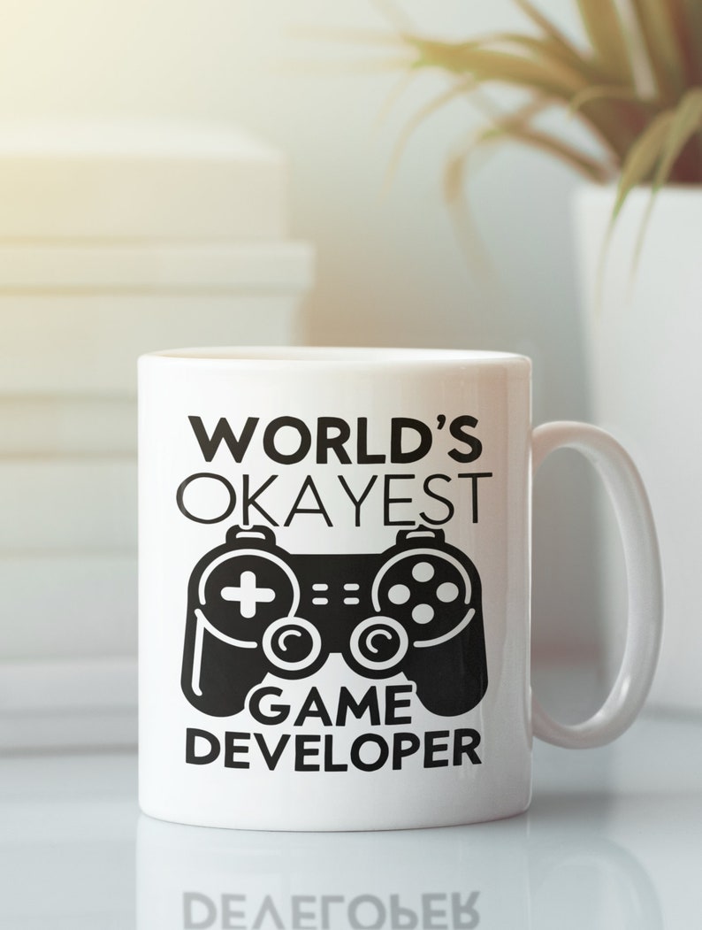 May include: White ceramic mug with a black design that says "World's Okayest Game Developer" with a black video game controller graphic.