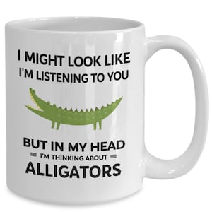 K&ouml;nnte beinhalten: Eine wei&szlig;e Keramik-Kaffeetasse mit einem schwarzen und gr&uuml;nen Alligator-Motiv und dem Text "Ich sehe vielleicht aus, als w&uuml;rde ich zuh&ouml;ren, aber in meinem Kopf = denke ich an = Alligatoren."
