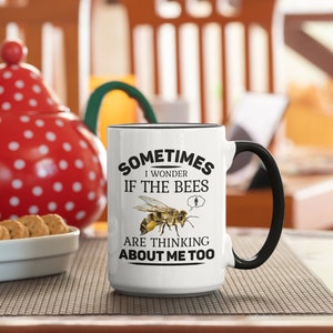 K&ouml;nnte beinhalten: Eine wei&szlig;e Keramiktasse mit schwarzem Henkel. Die Tasse hat einen schwarzen Text, der lautet "Sometimes I wonder if the bees are thinking about me too". Eine Cartoon-Biene ist mit einer Gedankenblase &uuml;ber ihrem Kopf abgebildet.