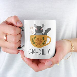 May include: White ceramic mug featuring a grey chinchilla sitting in a yellow teacup with a floral design. The text "Chai-Chilla" is printed on the mug.