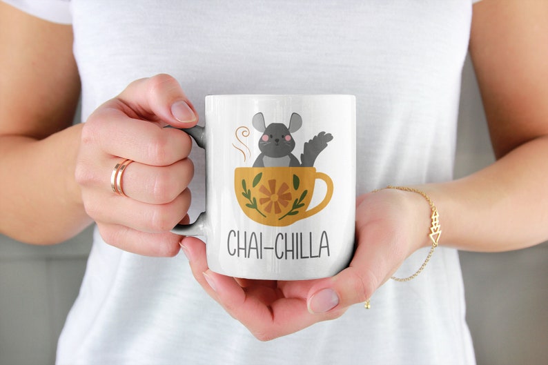 May include: White ceramic mug featuring a grey chinchilla sitting in a yellow teacup with a floral design. The text "Chai-Chilla" is printed on the mug.