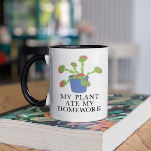Pode incluir: Caneca de cer&acirc;mica branca com al&ccedil;a preta. A caneca tem uma borda preta e apresenta uma ilustra&ccedil;&atilde;o de desenho animado de uma planta carn&iacute;vora em um vaso azul. O texto "MY PLANT ATE MY HOMEWORK" &eacute; impresso em preto abaixo da ilustra&ccedil;&atilde;o.