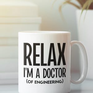 May include: White ceramic mug with the text "RELAX I'M A DOCTOR (OF ENGINEERING)" in black. The mug has a curved handle and is set against a blurred background with a stack of books and a plant.
