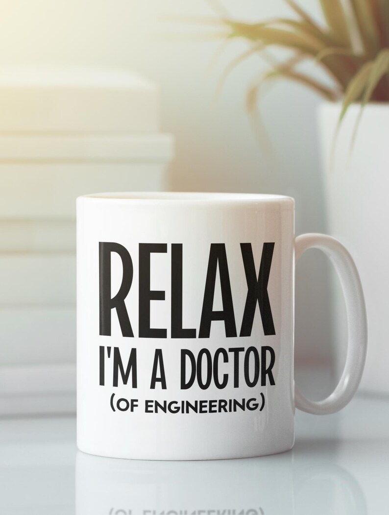 May include: White ceramic mug with the text "RELAX I'M A DOCTOR (OF ENGINEERING)" in black. The mug has a curved handle and is set against a blurred background with a stack of books and a plant.