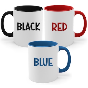 May include: Three white ceramic mugs with colored interiors and handles. One mug has a black interior and handle with the word "BLACK" in black. Another has a red interior and handle with "RED" in red. The third has a blue interior and handle with "BLUE" in blue.