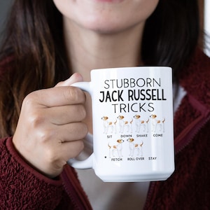 May include: White ceramic mug with the text "Stubborn Jack Russell Tricks" and illustrations of Jack Russell Terriers performing tricks. The tricks include sit, down, shake, come, fetch, roll over, and stay.