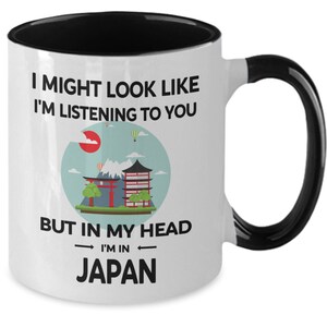 May include: White coffee mug with a black rim and handle. The mug has a graphic of a Japanese landscape with a red sun, mountains, and a pagoda. The text on the mug reads "I might look like I'm listening to you but in my head I'm in Japan."