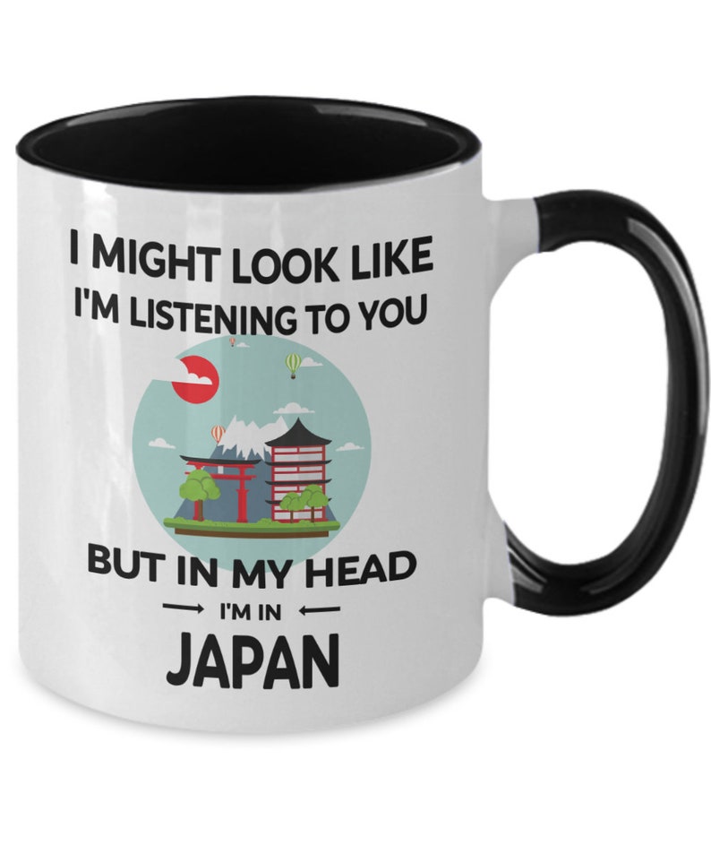 May include: White coffee mug with a black rim and handle. The mug has a graphic of a Japanese landscape with a red sun, mountains, and a pagoda. The text on the mug reads "I might look like I'm listening to you but in my head I'm in Japan."
