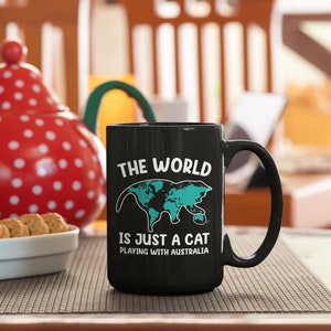 May include: Black ceramic mug with a white handle featuring the text "THE WORLD IS JUST A CAT PLAYING WITH AUSTRALIA." The design includes a teal-colored world map shaped like a cat. A red and white polka-dotted teapot and a bowl of cookies are in the background.