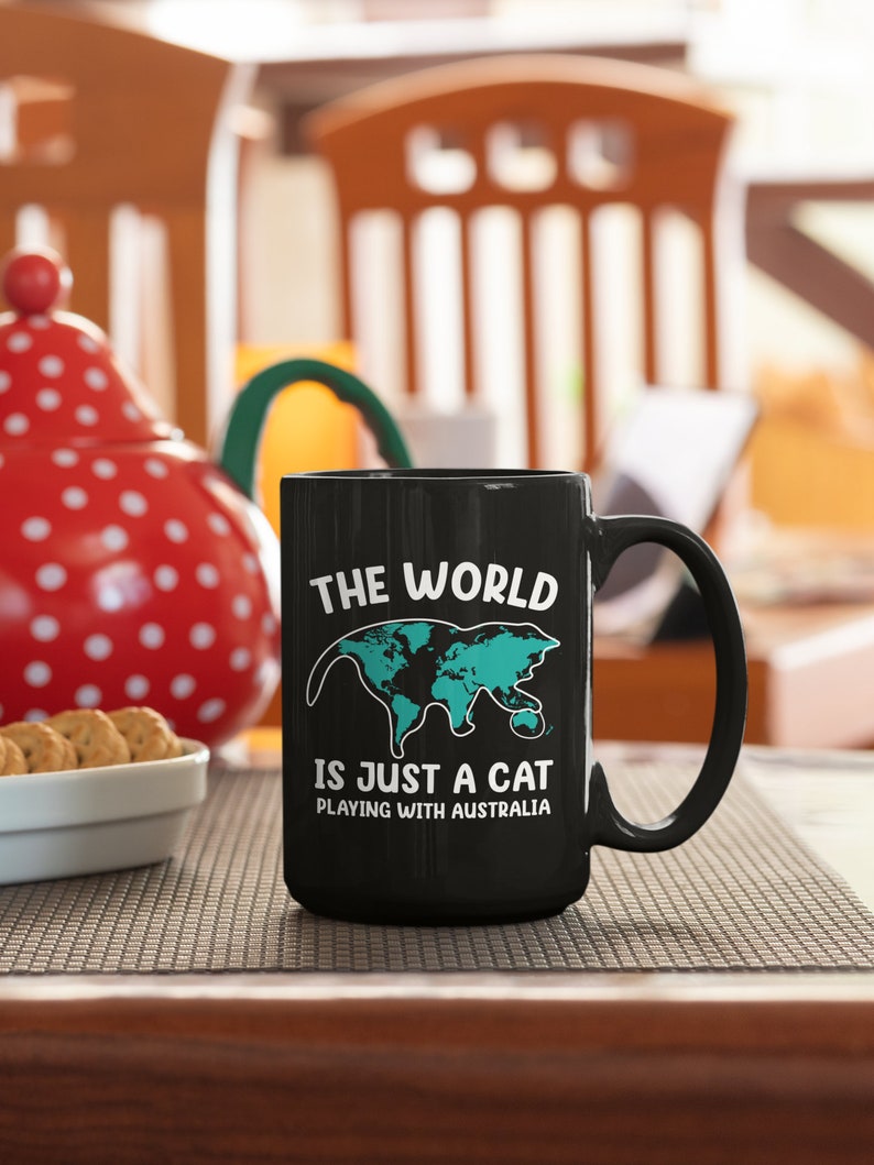 May include: Black ceramic mug with a white handle featuring the text "THE WORLD IS JUST A CAT PLAYING WITH AUSTRALIA." The design includes a teal-colored world map shaped like a cat. A red and white polka-dotted teapot and a bowl of cookies are in the background.