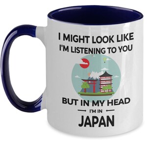 May include: White ceramic coffee mug with a blue rim and handle. The mug features a graphic of a Japanese landscape with a red temple and the text "I might look like I'm listening to you but in my head I'm in Japan."
