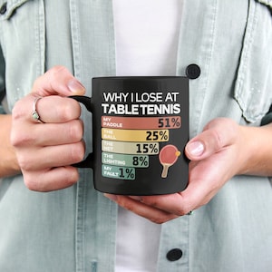 K&ouml;nnte beinhalten: Schwarze Kaffeetasse mit einer wei&szlig;en Grafik, die "Warum ich beim Tischtennis verliere" lautet, mit einer Liste von Gr&uuml;nden und Prozentangaben. Die Grafik enth&auml;lt eine kleine Illustration eines Tischtennis Schl&auml;gers und Balls.