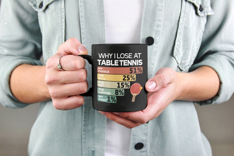 K&ouml;nnte beinhalten: Schwarze Kaffeetasse mit einer wei&szlig;en Grafik, die "Warum ich beim Tischtennis verliere" lautet, mit einer Liste von Gr&uuml;nden und Prozentangaben. Die Grafik enth&auml;lt eine kleine Illustration eines Tischtennis Schl&auml;gers und Balls.