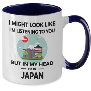 May include: White ceramic coffee mug with a blue rim and handle. The mug features a graphic of a Japanese cityscape with the text "I might look like I'm listening to you, but in my head I'm in Japan."