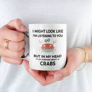 Pu&ograve; includere: Una tazza in ceramica bianca con un'illustrazione di granchio di cartone animato nero e rosso. La tazza recita "Potrei sembrare che ti stia ascoltando, ma nella mia testa sto pensando ai granchi."