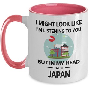 May include: White ceramic coffee mug with a pink rim and handle. The mug features a black and white graphic of a Japanese cityscape with the text "I might look like I'm listening to you, but in my head I'm in Japan."