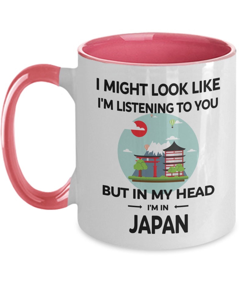 May include: White ceramic coffee mug with a pink rim and handle. The mug features a black and white graphic of a Japanese cityscape with the text "I might look like I'm listening to you, but in my head I'm in Japan."
