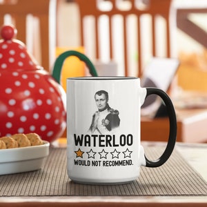 May include: White ceramic mug with a black handle. The mug features a black and white image of a man in a military uniform. The text "WATERLOO" is printed in black, followed by a one-star rating and the text "WOULD NOT RECOMMEND."