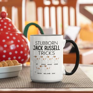 May include: White ceramic mug with a black handle and rim. The mug features the text "STUBBORN JACK RUSSELL TRICKS" with illustrations of Jack Russell Terriers demonstrating tricks like sit, down, and stay. A fun gift for dog lovers.