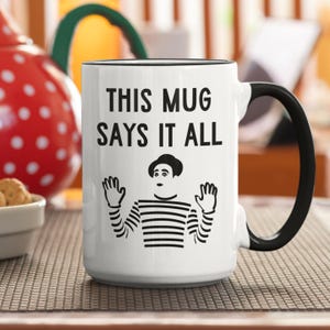 Op de afbeelding: Witte keramische mok met een zwarte rand en handvat. De mok heeft een zwart-witte illustratie van een mime met de tekst "This mug says it all".