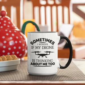 Op de afbeelding: Een witte keramische mok met een zwart handvat. De mok heeft zwarte tekst die luidt "Sometimes I wonder if my drone is thinking about me too."  Een zwart silhouet van een drone bevindt zich boven de tekst.