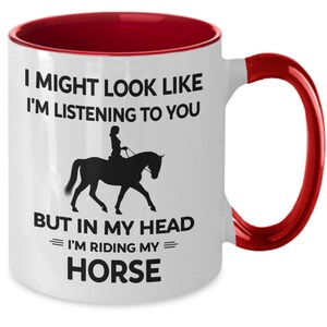 May include: White ceramic coffee mug with a red rim and handle. The mug features a black silhouette of a person riding a horse with the text "I might look like I'm listening to you, but in my head I'm riding my horse."