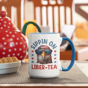 May include: White ceramic mug with a blue rim and handle. The mug features a cartoon bald eagle wearing sunglasses and a red, white, and blue bandana. The text "SIPPIN ON LIBER-TEA" is printed on the mug.