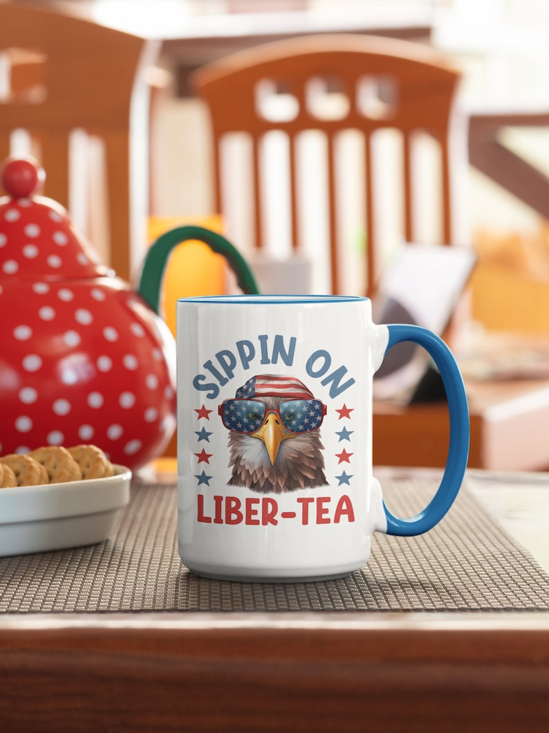 May include: White ceramic mug with a blue rim and handle. The mug features a cartoon bald eagle wearing sunglasses and a red, white, and blue bandana. The text "SIPPIN ON LIBER-TEA" is printed on the mug.