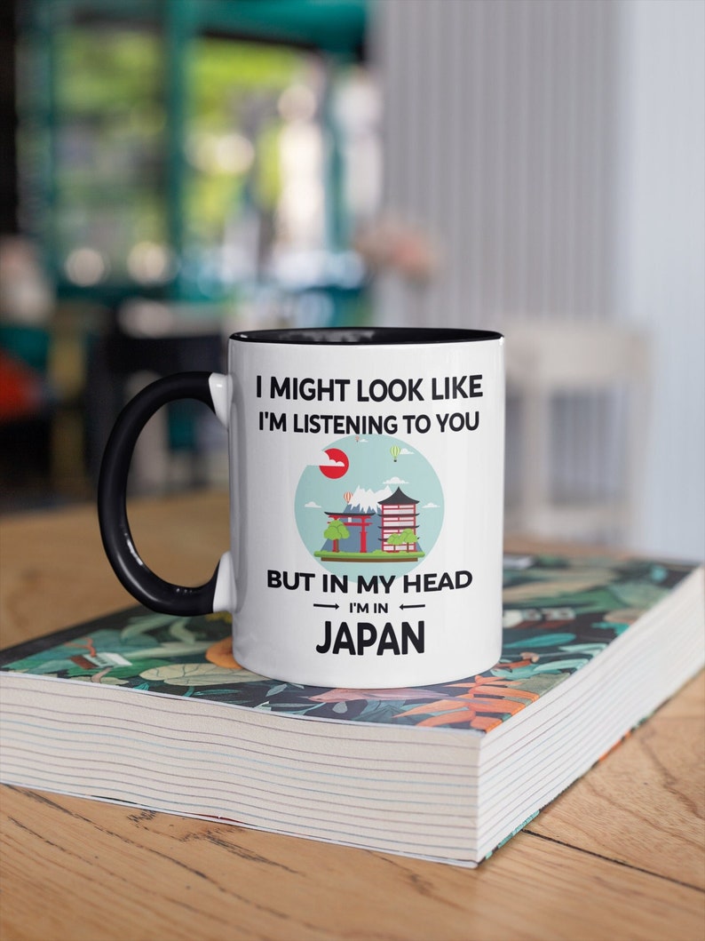 May include: White coffee mug with a black rim and handle. The mug has a black and white graphic with the text "I might look like I'm listening to you, but in my head I'm in Japan". The graphic includes a red sun, a mountain, a pagoda, and a hot air balloon.