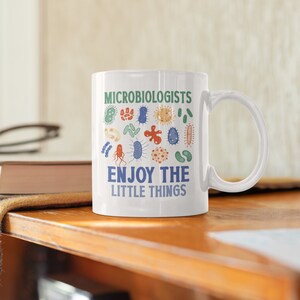 May include: White ceramic mug with a green and blue design of various microbes. The text "Microbiologists Enjoy The Little Things" is printed on the mug.