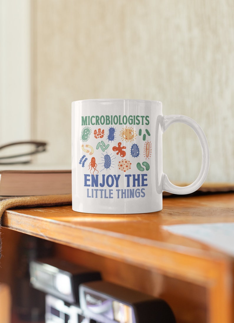 May include: White ceramic mug with a green and blue design of various microbes. The text "Microbiologists Enjoy The Little Things" is printed on the mug.