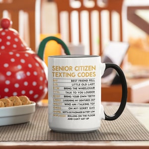 May include: White ceramic mug with a black handle and rim, featuring the text "SENIOR CITIZEN TEXTING CODES" in gold. The mug lists humorous acronyms and their meanings, such as "LOL" for "LITTLE OLD LADY".