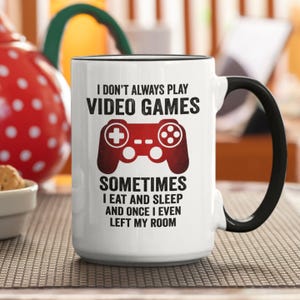 May include: White ceramic coffee mug with a black rim and handle. The mug features a red and black video game controller graphic and the text "I don't always play video games, sometimes I eat and sleep and once I even left my room."