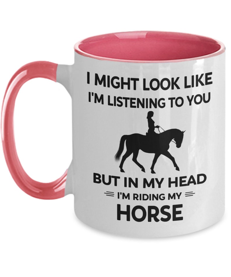 May include: White ceramic coffee mug with a pink rim and handle. The mug features a black silhouette of a person riding a horse with the text "I might look like I'm listening to you but in my head I'm riding my horse."