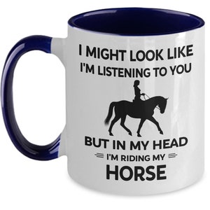 May include: White ceramic coffee mug with a blue rim and handle. The mug features a black silhouette of a person riding a horse with the text "I might look like I'm listening to you but in my head I'm riding my horse."