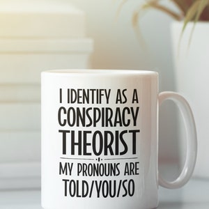 May include: White ceramic coffee mug with black text that reads "I identify as a conspiracy theorist. My pronouns are told/you/so."