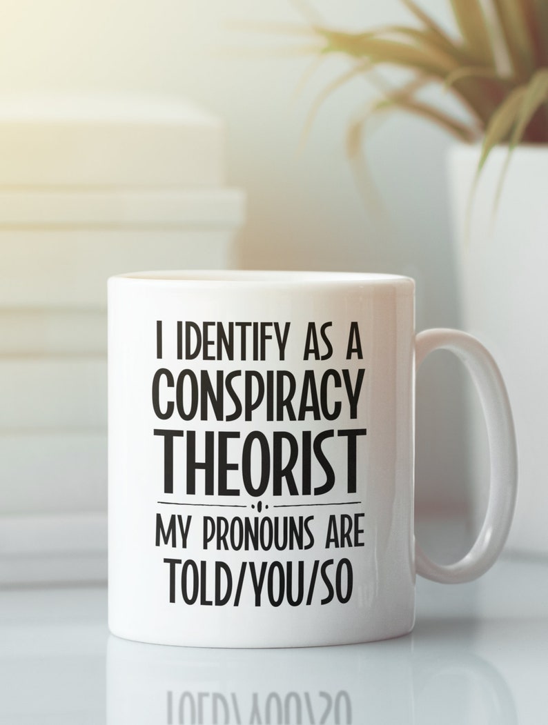 May include: White ceramic coffee mug with black text that reads "I identify as a conspiracy theorist. My pronouns are told/you/so."