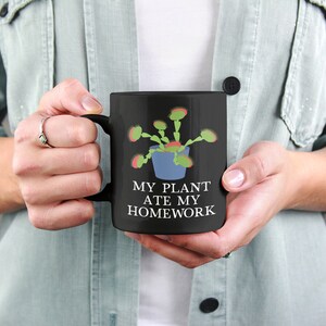 Pode incluir: Caneca de caf&eacute; preta com um design branco de uma planta carn&iacute;vora com o texto "My Plant Ate My Homework".