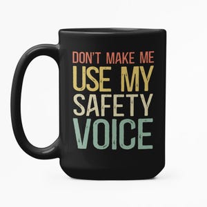 Op de afbeelding: Zwarte keramische mok met de tekst "DON'T MAKE ME USE MY SAFETY VOICE" in een retro kleurenschema. De mok heeft een groot handvat en een eenvoudig ontwerp, geschikt voor koffie, thee of andere dranken.