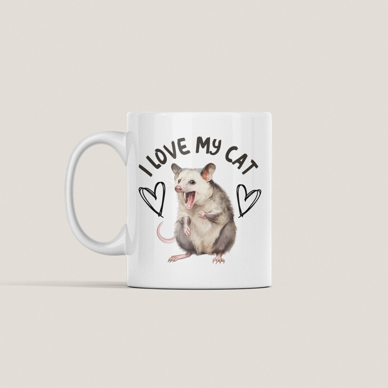 Puede incluir: Taza de cer&aacute;mica blanca con una ilustraci&oacute;n en blanco y negro de una zarig&uuml;eya con el texto "Me encanta mi gato" y dos corazones.