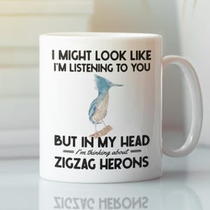 May include: White ceramic coffee mug with a black and blue illustration of a bird. The mug reads "I might look like I'm listening to you, but in my head I'm thinking about zigzag herons."