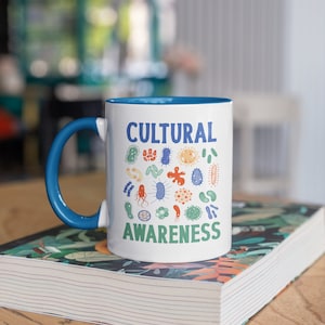 May include: A white ceramic mug with a blue rim and handle. The mug features a design of colourful germs with the text "CULTURAL AWARENESS" printed on it. The mug is resting on a book with a floral pattern.