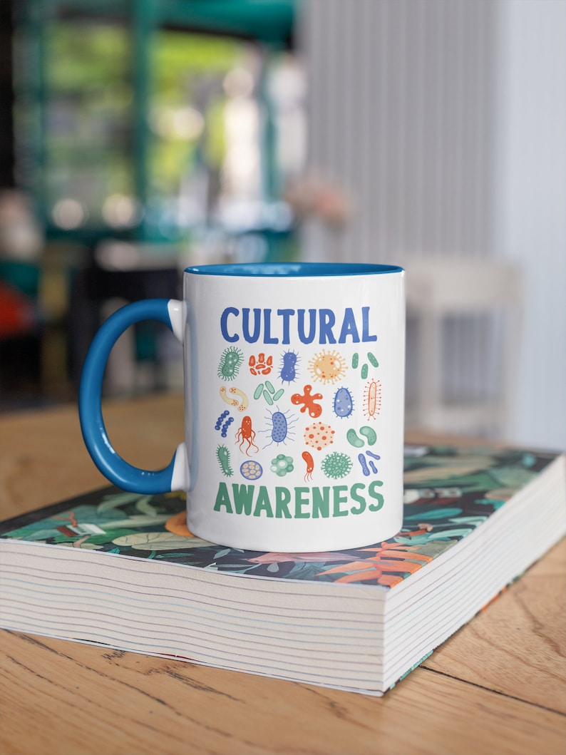 May include: A white ceramic mug with a blue rim and handle. The mug features a design of colourful germs with the text "CULTURAL AWARENESS" printed on it. The mug is resting on a book with a floral pattern.