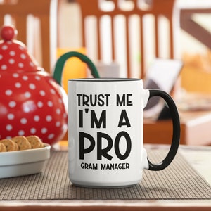 May include: White ceramic mug with a black handle and black text that reads "Trust me I'm a pro Gram Manager".