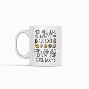 Op de afbeelding: Witte keramische mok met een zwarte afbeelding met de tekst "Not all who wander are lost. Some are just looking for cool rocks." De afbeelding bevat een rij met verschillende gekleurde stenen.