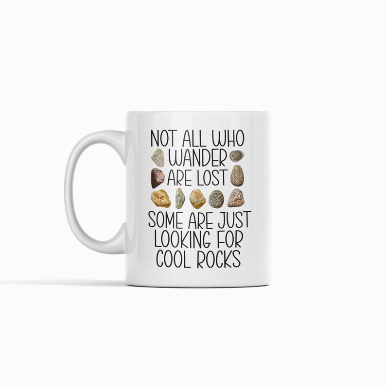 Op de afbeelding: Witte keramische mok met een zwarte afbeelding met de tekst "Not all who wander are lost. Some are just looking for cool rocks." De afbeelding bevat een rij met verschillende gekleurde stenen.